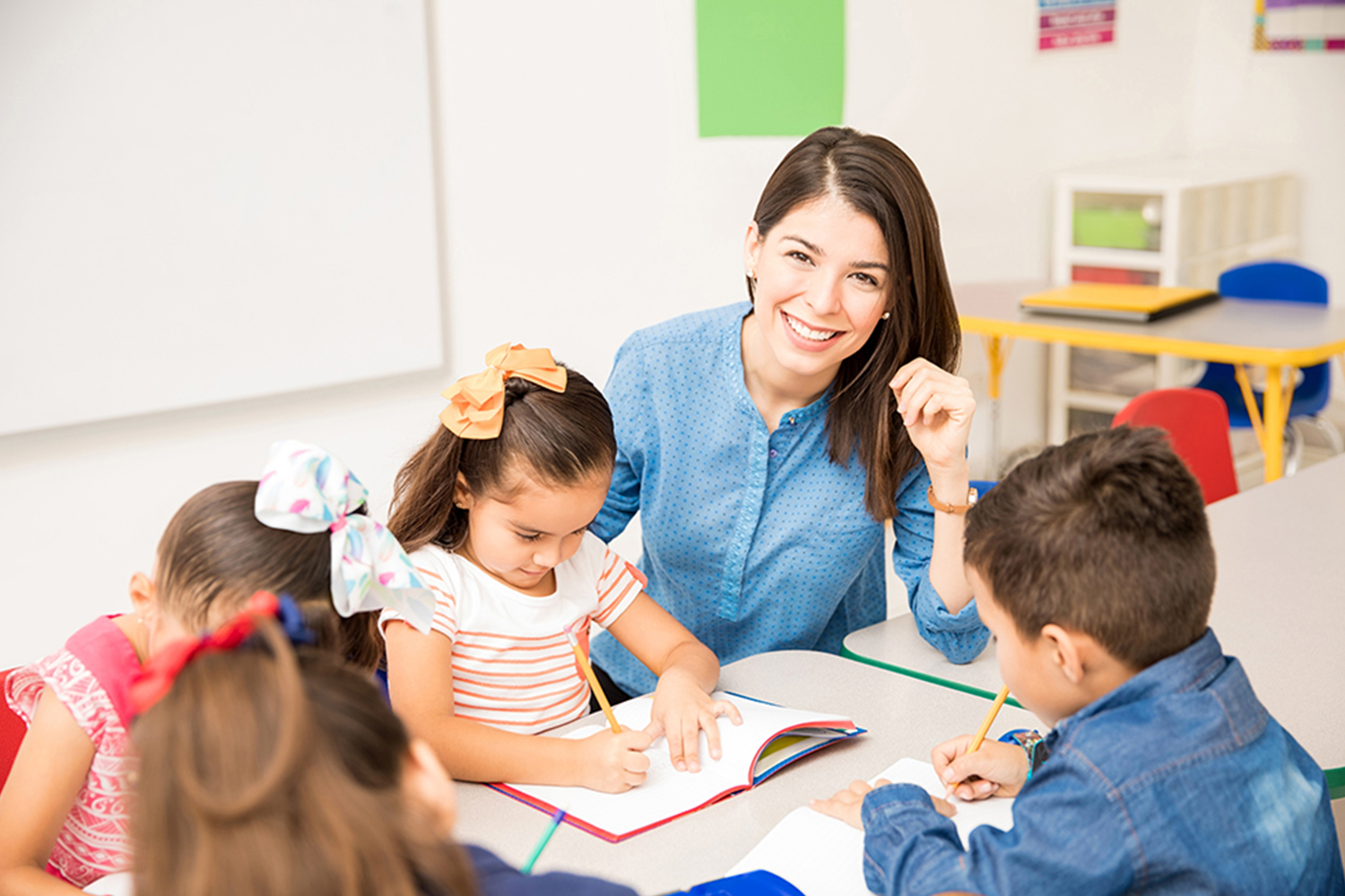 A female teacher with a blue shirt is in a classroom, smiling, as her young students work on a project.